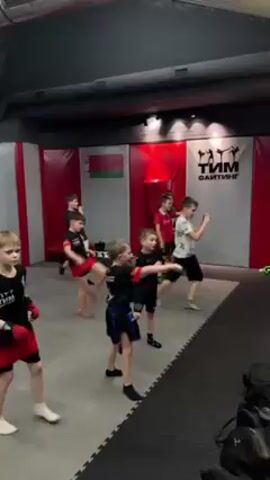 Several children, approximately 8-10 years old, are practicing martial arts kicks in a gymnasium. They are barefoot and wearing shorts with martial arts logos. Some have boxing gloves. The gym has padded walls with red and white accents. A Belarusian flag is visible on the wall.  There is a logo that includes the words "ФАЙТИНГ." The children are spaced apart and kicking in unison.  The floor is gray with a textured pattern. A black bag rests in the foreground.