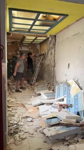 The video shows a room undergoing demolition. Two men are present. One man, wearing glasses and a gray t-shirt and shorts, stands on the floor to the left, observing. The other man, wearing a dark t-shirt and shorts, stands on a ladder to the right, using a demolition tool to break apart the wall and ceiling. Debris, including plaster and what appears to be wallpaper with a blue and white star pattern, is scattered across the floor. The room is in disarray, with exposed walls and ceiling elements. The ceiling has a yellow border and a grid-like structure.