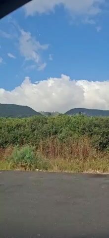 The video displays a time-lapse view from inside a vehicle, moving along a paved road. On the right side of the road, there is a metal guardrail. Beyond the road and guardrail, a dense green hillside is visible, covered in lush vegetation and some dry grasses at the base. In the background, rolling hills form the horizon, and a bright blue sky with scattered white clouds fills the upper portion of the frame. The camera angle suggests the viewer is looking out the side window of the vehicle.