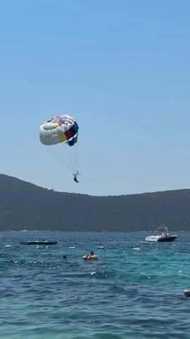 A person is parasailing over a body of water. A multicolored parachute is visible above them. The water is blue with some waves. In the distance, there are hills covered in trees and a boat. Some people are swimming or floating in the water. The sky is clear and blue.