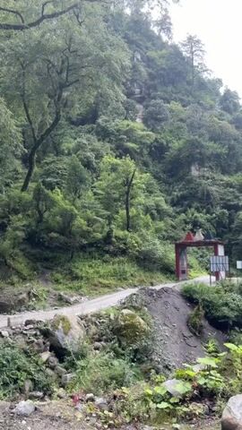 The video pans from right to left, showing a lush, mountainous landscape. A dirt road curves through the scene, bordered by rocks and dense green foliage. Trees with thick trunks and abundant leaves dominate the upper portion of the frame. In the foreground, a small waterfall cascades over moss-covered rocks, feeding into a stream. The sky appears overcast. Further up the mountainside, a structure resembling a traditional gateway or archway is visible. The overall impression is one of a natural, somewhat wild environment.