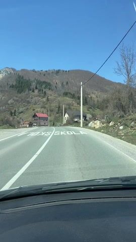 The video shows a drive-through of a road in a mountainous region on a clear, sunny day. The road is paved with a white line dividing the two lanes. The view is from inside a car, looking through the windshield. There are houses and utility poles along the side of the road. In the distance, there's a mountain range, and some snow can be seen on the peaks. A road marking that reads “PAZI! SKOLA" is briefly visible.