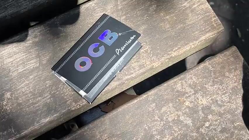 A close-up, high-angle shot shows a pack of OCB rolling papers with holographic lettering on a weathered wooden surface. Sunlight casts shadows across the pack and the wooden planks. The pack is partially open, revealing a small white object inside. A hand is visible in the top right corner of the frame, reaching towards the pack. The environment appears to be outdoors, with some dry leaves scattered on the wood.