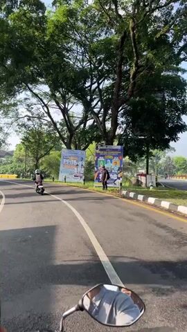 The video shows a first-person view from a motorcycle traveling on a paved road lined with trees. There are other motorcycles on the road, traveling in the same direction. On the side of the road, there are two large posters, one with a person in a suit and the other with a group of people with backpacks. A man stands next to the second poster. The road has a yellow guardrail. The sky is blue and clear. Houses and other buildings can be seen in the distance.