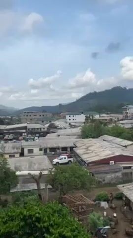 The video presents a high-angle view of a rural residential area. The scene encompasses single-story buildings with corrugated metal roofs, mostly in neutral tones. A dirt road runs between the buildings. In the background, a lush, green hillside rises, partially obscured by clouds in the sky. The weather appears partly cloudy, with a mix of blue sky and scattered white clouds.