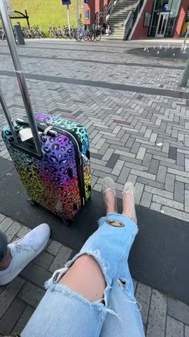 The video shows a person's lower body sitting on the ground next to a colorful leopard-print suitcase. The person is wearing light blue ripped jeans and beige ballet flats. Next to them, a person is sitting with a white Adidas sneaker visible. The background shows a brick-paved area, a set of stairs leading to a building, bicycles parked, and some greenery in the distance. The scene appears to be outdoors, possibly at a train station or public transportation area.