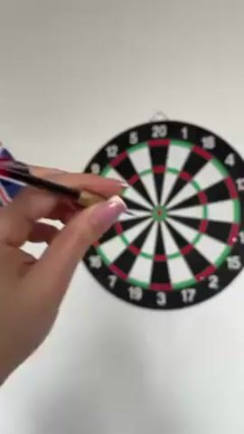 A hand with manicured fingernails throws a dart at a dartboard on a white wall, and the dart hits the outer edge of the single 20 area.