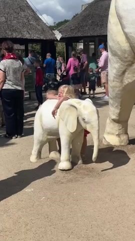A blonde-haired girl is sitting on a white elephant statue with her arms wrapped around its neck. She is looking directly at the camera with her mouth open, seemingly yelling or making a face. Another child, partially obscured, is behind her, also on the statue. The elephant statue is placed on a dirt path, with other people in the background walking and standing near thatched-roof structures. The scene appears to be outdoors, possibly at a zoo or park, with sunlight casting shadows on the ground. The lighting is bright, suggesting daytime.