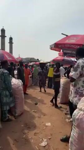 The video shows a busy outdoor market scene. The ground is dirt, and there is litter scattered around. Large sacks of goods are stacked along the side of the path. People are walking through the market, some carrying goods on their heads. There are umbrellas overhead to provide shade. In the background, the minaret of a mosque is visible.