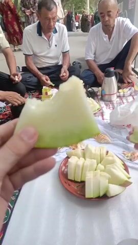 A person's hand holds a slice of honeydew melon with a bite taken out of it. In the background, two older men sit on the ground, facing each other. They are dressed casually. One man wears a white polo shirt and dark pants, while the other wears a white shirt and dark pants. Both men are eating. There is a platter of sliced honeydew melon in front of them, along with some flatbread. A thermos and other items are also on the ground. In the further background, other people are walking. The setting appears to be outdoors, possibly a park or public gathering area, with trees and pavement visible.