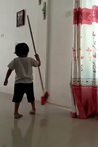 A young child stands in a room and swings a broom. The child is barefoot and wears a grey shirt and dark shorts. The child is facing away from the camera. The broom has a long metal handle and a red and grey brush. The room has a shiny tiled floor, white walls, and a patterned curtain in the background. There is a framed picture hanging on the wall. The child appears to be playing with the broom, rather than actually sweeping.