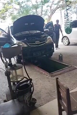 A black Toyota Avanza is parked over a vehicle inspection pit. A person, wearing a black t-shirt with a yellow and black graphic logo on the back, is partially visible inside the pit, appearing to work on the underside of the car. The license plate of the car reads "BL 1674 NP". To the left of the car, there is a piece of automotive maintenance equipment with a transparent container filled with liquid and a hose. In the background, another person is walking away from the car, and to the right, a green coiled hose hangs from a structure. The environment appears to be an open-air garage or workshop.