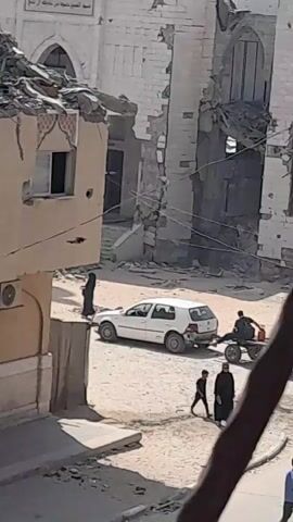 A group of people are seen in a desolate, war-torn environment. A white car is parked in the middle of a street, which appears to be made of dirt and rubble. A cart pulled by two people is in motion, carrying a large red container. One person is seated on the cart, while another walks beside it. In the background, several buildings show significant damage, with collapsed walls and debris scattered around. The overall scene suggests a post-conflict or active conflict zone where life is being carried on amidst destruction.