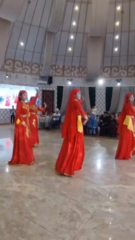 A group of young women in matching red dresses with gold trim and yellow sashes are performing a synchronized dance in a large, well-lit room with a domed ceiling. The floor is polished marble. In the background, there are people seated at tables and a large screen displaying an image of dancers. The dancers are mostly facing away from the camera, except for one in the foreground who is facing the camera with her arms extended.