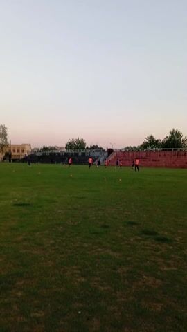 The video shows a grassy sports field with several people practicing soccer. Some people are wearing orange vests. A set of stadium-style concrete bleachers are in the background. Trees, buildings, and a pale sky are also visible. The camera appears to be hand-held and moves erratically.