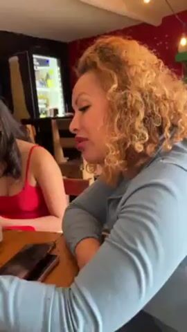 The video shows three women sitting around a wooden table in a bar or restaurant. The woman closest to the camera has curly, light brown hair and is wearing a light blue sweatshirt. The woman to her left is wearing a red tank top and has long, dark hair. A third woman, wearing a grey sweatshirt, is also visible. Each woman has a phone on the table in front of them and appear to be looking at them. The table also has beer bottles and glasses on it. The background shows a bar with shelves of drinks, a television, and musical decorations. There are overhead lights and sunlight coming in. The restaurant appears to have dark colored walls, with the exception of a red section. The scene is brightly lit.