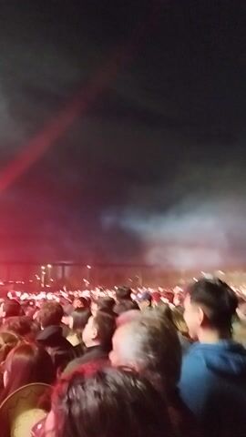 The video captures an energetic night scene of a live concert or festival. A large crowd of people is gathered, their faces partially visible in the foreground, looking towards a brightly lit stage. The atmosphere is vibrant, with a significant amount of stage fog or smoke contributing to the visual effects.

The stage itself is large and features professional lighting rigs with an array of white and blue lights, as well as several large screens displaying blue and red graphics. Performers, including a drummer and guitarists, are visible on stage, engaged in their act. Red and blue stage lights illuminate the scene, casting dynamic colors across the fog. The overall impression is one of a lively public event taking place at night, likely in an open area like a park or plaza in Maipú, Chile. The crowd appears engaged and is enjoying the performance.