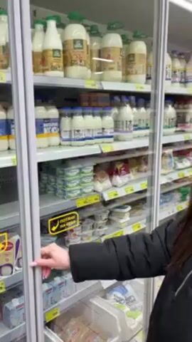A woman wearing a black jacket and black beanie opens the door to a refrigerated section in a grocery store. The refrigerated section is stocked with milk, yogurt, and other dairy products. She looks into the refrigerator, seemingly to make a selection. The shelves are white, and the door frame is grey. She has brown hair and makeup and is looking directly toward the refrigerated section. The store has bright, overhead lighting.