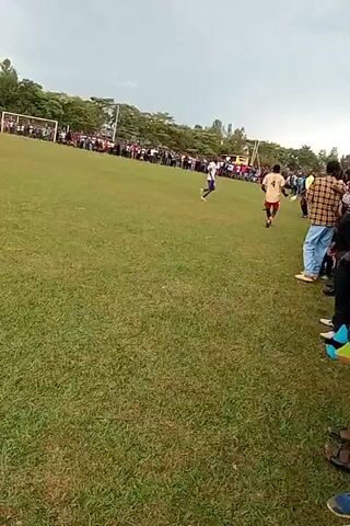 The video shows a soccer game. There are several people on the field playing soccer. One person is wearing a yellow shirt and red shorts and is running with the ball. Another person is wearing a blue and white uniform and is also running. There is a crowd of people standing on the sidelines watching the game. There is a goal in the distance. The sky is overcast.