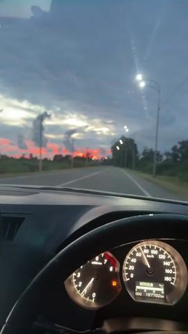 The video shows a first-person perspective from inside a car driving on a paved road. The time is dusk, and the sky is filled with dramatic clouds tinged with red and orange from the setting sun. Streetlights are visible along the side of the road, and trees and fields border the road. The car's dashboard is visible, including the steering wheel and the speedometer, which shows the car traveling at approximately 80 km/h. Two other vehicles, a dark van and a dark sedan, are seen on the road ahead. The camera appears to be mounted on the dashboard or windshield.