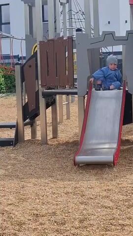 A baby wearing a blue jumpsuit and a gray beanie sits at the top of a metal slide on a playground. The playground has wood chip ground cover and a gray and brown play structure resembling a castle. The baby begins to slide down the slide, their feet touching the surface. The baby reaches the bottom of the slide.