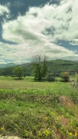 The video shows a landscape of rolling green hills under a cloudy sky, captured from a moving vehicle. The foreground contains lush green grass and patches of brown soil, with scattered trees dotting the landscape. In the background, the hills are covered in dense forest, creating a sense of depth. The sky is filled with large, fluffy clouds, and patches of blue are visible between them. The overall impression is of a peaceful, rural scene.
