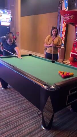 The scene shows a man and a woman playing a game of pool in a brightly lit game room. The man is on the left, wearing glasses and a dark shirt, with his sleeves rolled up. He has just hit the cue ball, scattering the red and yellow pool balls across the green felt of the pool table. The woman, standing to his right, wears a yellow shirt and jeans, holding a pool cue while watching. A red, white, and blue arcade game stands nearby, and another arcade game, featuring bright lights, is visible in the background. The floor is covered with a colorful striped carpet. The pool table itself has a dark brown frame with chrome corner accents.