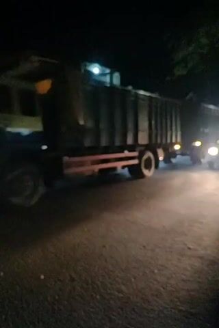 The video is a shaky, blurry nighttime recording of a busy street. A large, dark-colored truck with a green trailer is the main subject, driving away from the camera and to the left. Its headlights are on, illuminating the asphalt road. Several motorcycles are also on the road, some passing the truck and others driving alongside it. One motorcycle in the foreground is particularly bright, its headlight shining directly at the camera. Buildings and trees are visible in the background, partially obscured by darkness and the motion of the camera. The overall impression is one of chaotic night traffic.