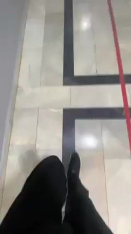 The camera moves through a school hallway. The floor is covered in light-colored marble tile with black marble borders. A red tape line follows the floor, providing direction. The walls are painted grey on the lower half, white above.  Pictures hang on the walls. The door to a "Tarikh Xonasi" is visible. A person wearing black sneakers walks forward.
