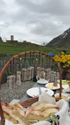 The video shows a close-up view of a breakfast spread laid out outdoors, with a blurred mountain landscape in the background. The foreground is dominated by a wicker basket filled with freshly baked bread. Behind the basket, on a table, are plates, cutlery, small jars of preserves or condiments, and slices of butter. A decorative element featuring miniature stone towers suspended from a curved wooden frame hangs over the table. The backdrop reveals a green, hilly terrain dotted with buildings, a wooden fence, and a snow-capped mountain under a cloudy sky. Yellow flowers in a pot add a splash of color to the right side of the frame. The overall scene suggests a rustic, outdoor dining experience in a mountainous region.