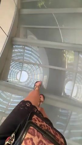 The video shows a first-person perspective of someone walking on a glass walkway that curves around a large, multi-level atrium with a circular water feature in the center. The walkway has a metal railing. Below the walkway, on different levels of the atrium, are shops and people. The water feature has glowing green accents and a rock formation in the center. The person walking is wearing sandals and carrying a leopard-print bag. The overall impression is that of a modern shopping mall or public space with an impressive architectural design.