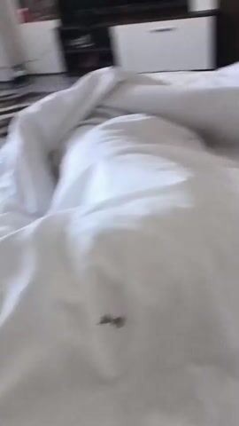 The video opens with a close-up shot of a white, crumpled duvet or bed sheet. A small, dark, fly-like insect is resting motionless on the fabric in the foreground. The background, which is out of focus, reveals an indoor room, likely a bedroom, with furniture including a dark wardrobe or shelf unit and a white cabinet.

Around the 0:02 mark, a human hand enters the frame from the bottom left, reaching towards the insect. A finger gently nudges the insect at 0:03, indicating it might be stunned or deceased, as it doesn't react. The finger continues to push the insect, causing it to tumble and slide across the white linen before falling off the edge of the duvet and out of frame by 0:05. The hand then retracts. The video concludes with a shot of the now-clear white duvet, with the blurred background of the room still visible.

The scene takes place indoors, likely a bedroom, during what appears to be daytime due to the ambient brightness. There is no visible text. The activity involves a person gently removing a small insect from their bed linen. The location context provided is Boliartsi, Bulgaria.