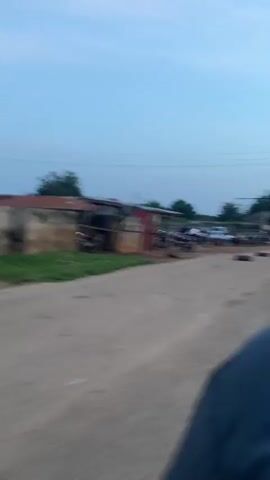 The video shows an outdoor scene filmed from a moving vehicle on an unpaved road. Two dark tires lie on the dirt road in the foreground. To the left, simple structures with corrugated metal roofing, one red and one gray, are visible. A large group of people is gathered in the middle ground, with numerous motorcycles parked among them and a red car in the background. Several individuals are walking on the right side of the road. Power lines span across the clear, light blue sky.
