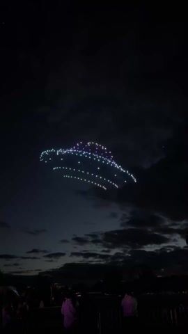 At night, a group of drones is flying in formation. The drones, which are lit in white, green, and purple, are arranged in an abstract shape in the sky. The sky is dark with scattered clouds and the drones cast light into their surrounding air. In the foreground, a crowd of people is gathered watching the display. They are silhouetted against the dark ground. Music is audible.