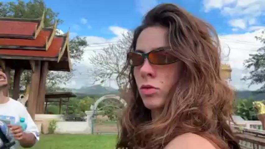 The video shows a woman in sunglasses in the foreground with a man in the background. The woman has long brown hair and is looking off to the side. The man has a mustache and a bandana. He is holding a water bottle and has a camera around his neck. In the background, there is a Buddhist temple and a green lawn. The sky is partly cloudy. The camera is moving slightly, and there is indistinct chatter in the background.