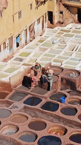 Captured from an elevated perspective, the video displays three men actively engaged in the tanning process at what appears to be a traditional tannery. The tannery is characterized by numerous circular and square pits or vats arranged in a grid pattern. These vats are filled with liquids of varying colors, including dark browns and blues. Some of the vats are empty or contain minimal fluid, revealing their earthen construction. Above this area, a series of square vats are laid out in a similar grid, filled with a lighter colored liquid. The men stand alongside the vats, handling what appears to be animal hides or skins. They are moving and soaking the hides, working together to move them from one vat to another. The surrounding architecture includes multi-story buildings with windows and doors, suggesting an urban setting. The lighting indicates a sunny day.