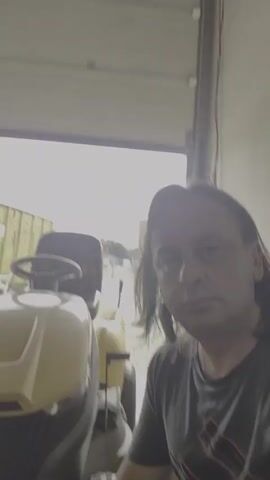 This is a short video clip, likely a selfie, featuring a person with long, dark hair and a gray t-shirt. They are positioned in the foreground on the right side of the frame. Behind them, to the left, is a yellow lawn tractor. Further in the background, through an open garage door, a bright, possibly overcast sky is visible, with some structures and yellow shipping containers outside.

The scene appears to be a workshop or garage, with industrial-looking elements such as the roller door and exposed beams in the ceiling. The lighting suggests it is daytime. The person is looking directly at the camera with a neutral expression.

A notable detail is the text visible on the side of a yellow vehicle in the background, which appears to be a forklift or similar industrial equipment. The text, in reverse due to being on the vehicle, reads: "9000 HINRICH Miel-Service 0800 SSS 28 28 88". This suggests a service company name and a phone number. The location context of Papenburg, Germany, would align with the German text.

The overall impression is a casual snapshot taken in a work environment, possibly during a break. There is no discernible activity beyond the recording of the video itself.