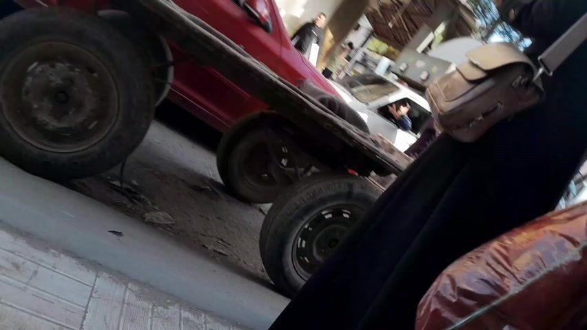 The video shows a horse-drawn cart traveling on a street. A red car is parked nearby.  Several people are present, some watching the cart, and one appears to be holding the horse's reins. The environment looks like a street in a populated area, with buildings and trees visible in the background. The perspective is from someone following closely, potentially walking alongside the cart.