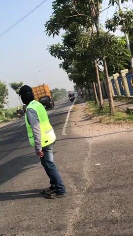 The video shows a person wearing a black mask and a bright yellow-green vest standing on the side of a paved road. The person is wearing blue jeans and what appear to be sandals. In the background, there is a large yellow truck and several motorcycles. Trees line the side of the road, and there is a blue and yellow fence or wall. The sky is clear and blue, suggesting it is daytime. The road appears to be a two-lane road with traffic moving in both directions. The person is facing away from the camera and appears to be looking towards the traffic.
