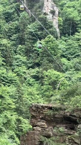The video shows two cable cars suspended on wires between lush green hills and rock formations. One cable car is yellow and the other is green. The surrounding landscape is dominated by dense forest and jagged rock faces. The weather appears overcast. Sounds of conversations in the background can be heard.