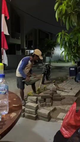 A man is working at night to lay paving stones. He is wearing a hat, a long-sleeved shirt, and shorts. He is holding a shovel and is in the process of placing a paving stone. There are stacks of paving stones and a pile of sand next to him. In the background, there is a motorcycle and a building. There are also Indonesian flags visible.