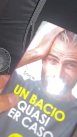 The video shows a close-up of a book cover titled "UN BACIO QUASI PER CASO" by Nicole Snow. The cover features a man with his hands on his head, looking at the camera. The book is held by a person whose thumb is visible on the left side of the frame. The video then transitions to a blurry, close-up shot of a young man with blondish-brown hair and a beard, lying down and looking at the camera. He is shirtless and wearing a silver necklace with a cross pendant. The background appears to be a bed or couch with dark fabric.