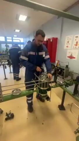 A medium-shot video shows a man wearing a navy blue jumpsuit with white stripes standing in a workshop and working on a large piece of machinery. He is holding a small tool and appears to be tightening or adjusting something on the top of the machine. He has short dark hair, a beard, and is wearing glasses. The machine is mostly silver and green and has various pipes and connections. Behind him, another person in a similar jumpsuit is working on a different machine. The room has bright fluorescent lighting and appears to be a training or educational facility, with various pieces of equipment and instructional posters on the walls. There are red lockers visible in the background. The camera moves slightly to follow the man's actions as he works on the machinery.