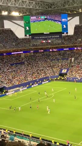 This is a high-angle, wide shot from a packed stadium during a soccer match. The field is green with white lines and goals at either end. There are numerous players in black and white uniforms on the field. A large scoreboard above the field displays the score as 1-1, the FIFA World Cup 2026 logo, and a smaller screen showing a replay of the game. The stadium seats are filled with a large crowd wearing various colored clothing, some holding their arms up. The overall atmosphere appears to be lively and engaged with the game.