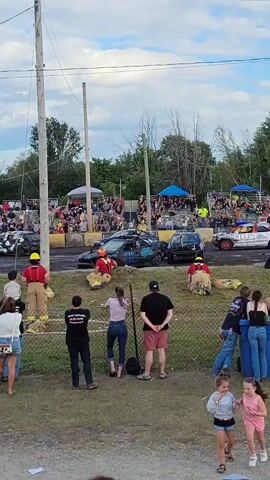 A demolition derby is underway. Several vehicles, some damaged, are in the arena. Spectators are seated in bleachers in the background. A group of people in firefighting gear, some wearing yellow hard hats, are standing near the fence, appearing to be on standby. There are also several spectators standing in the foreground, observing the event. The sky is cloudy.