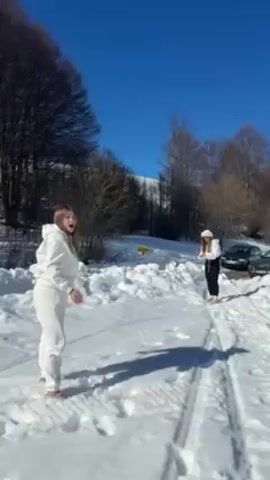 Two young women are having a snowball fight in a snowy landscape on a sunny day. One woman is wearing a white hoodie and sweatpants, and the other is wearing a black and white outfit with a white hat. They are standing on a snow-covered road, surrounded by snowdrifts and trees. In the background, there are a few cars parked. The woman in the white outfit throws a snowball at the woman in black and white.