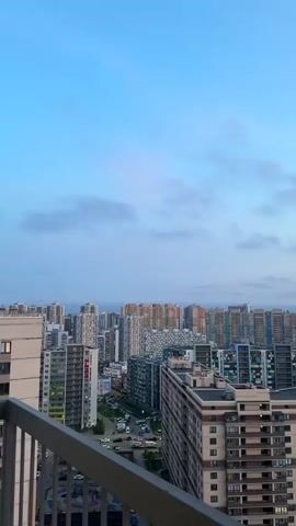 The video shows an urban panorama, likely from a high vantage point like a balcony, overlooking a densely populated area filled with multi-story residential buildings. The buildings vary in design and color. Roads with moving cars are visible between the buildings, indicating active urban life. The sky is partly cloudy with a blue hue, suggesting either early morning or late evening. The camera is stationary, capturing a static view of the cityscape and the sky.