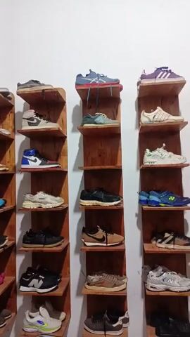 The video captures an indoor scene displaying a collection of athletic shoes. Multiple pairs of sneakers, encompassing various brands such as Nike, Adidas, New Balance, Hoka, and Asics, are neatly arranged on individual wooden shelves. These shelves are mounted vertically on a plain white wall, forming several tall, narrow display units. The shoes come in a wide array of colors and styles, suggesting a diverse collection. A small portion of a blue chair is visible in the bottom right corner of the frame. The overall setting appears to be a dedicated display area for shoes, possibly within a store or a personal collection space.