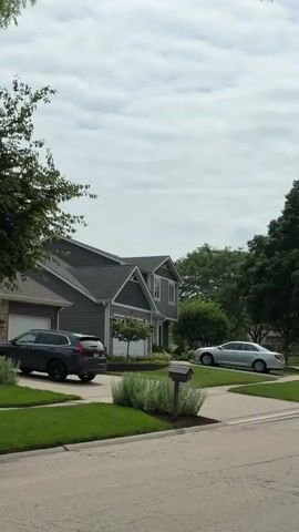 This video shows a panning view of a suburban street. There are houses, cars, lawns, and trees. The sky is overcast. In the foreground, there is a sidewalk curving around a patch of grass. A mailbox is visible near the sidewalk. A silver sedan is parked on a lawn to the right. A gray SUV and a white car are parked in a driveway to the left. The video pans right and then moves forward, showing more houses and trees along the street. The overall impression is of a quiet, residential neighborhood.