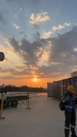 The video shows a sunset scene with a person in the foreground wearing a yellow hard hat and a blue safety vest, looking at their phone. In the background, there are shipping containers and a few other people also wearing safety vests. The sky is filled with clouds and sun rays are visible.